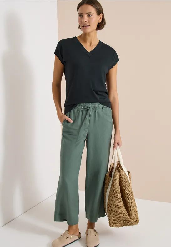 Eine Frau trägt ein schwarzes T-Shirt, locker geschnittene grüne Hose und hält eine große geflochtene Tasche.