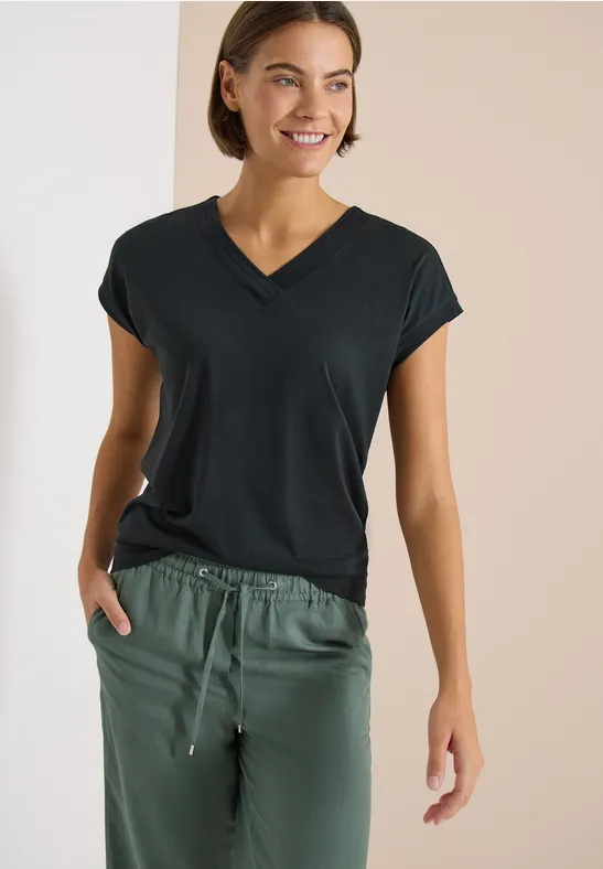 Frau in schwarzem V-Ausschnitt-T-Shirt und grüner Jogginghose, Hand in Tasche, vor neutralem Hintergrund.