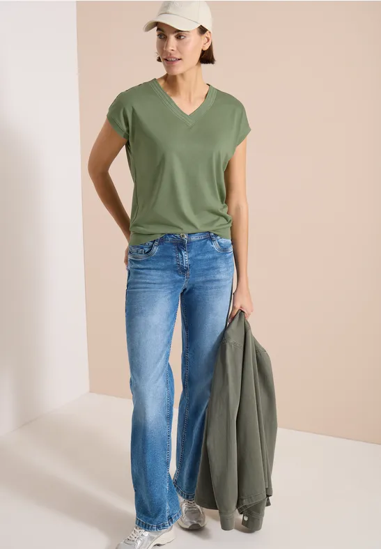Frau in grünem V-Shirt, blauen Jeans, hält olivegrüne Jacke vor hellem Hintergrund.