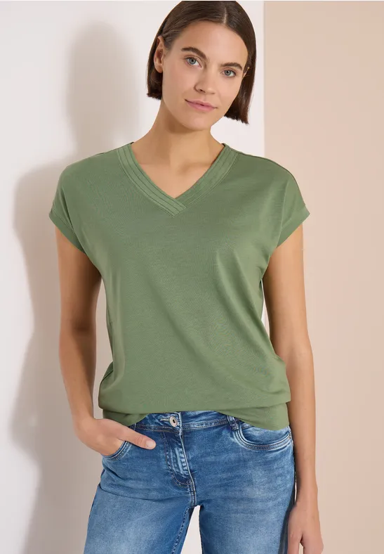 Frau trägt grünes V-Shirt und blaue Jeans vor hellem Hintergrund.
