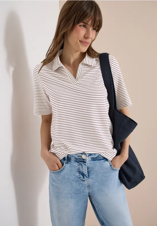 Frau in gestreiftem Kurzarm-Polo, hellblauen Jeans und schwarzer Umhängetasche vor schlichtem Hintergrund.