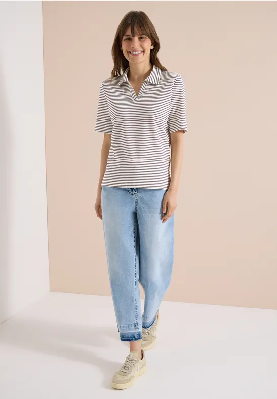 Frau in grauem Poloshirt, lockeren hellblauen Jeans und beigen Sneakern vor neutralem Hintergrund.