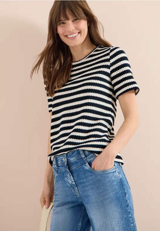 Frau trägt schwarz-weiß gestreiftes T-Shirt und blaue Jeans, steht vor neutralem Hintergrund.