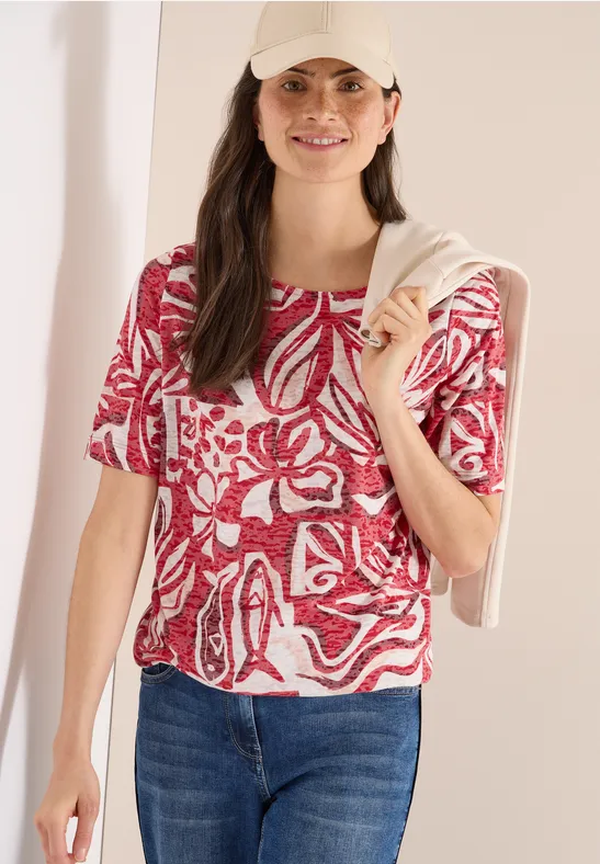 Frau in rotem, floral gemustertem Top, blauer Jeans und einer beigen Strickjacke, lässig über die Schultern gelegt.