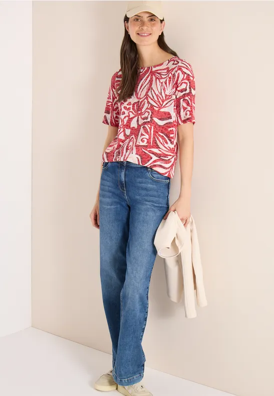 Eine Person trägt eine rote gemusterte Bluse, weite Jeans und hält eine helle Jacke in der Hand.