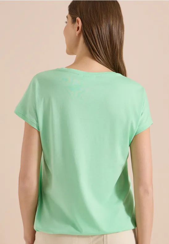 Frau von hinten in schlichtem, hellem grünen T-Shirt vor neutralem Hintergrund.