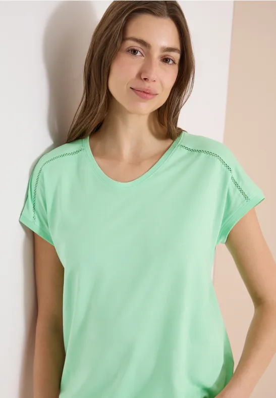 Junge Frau trägt mintgrünes T-Shirt und lehnt an hellem Hintergrund.