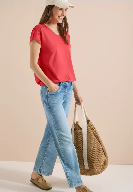 Frau in rotem T-Shirt, blauen Jeans und großer geflochtener Tasche vor neutralem Hintergrund.