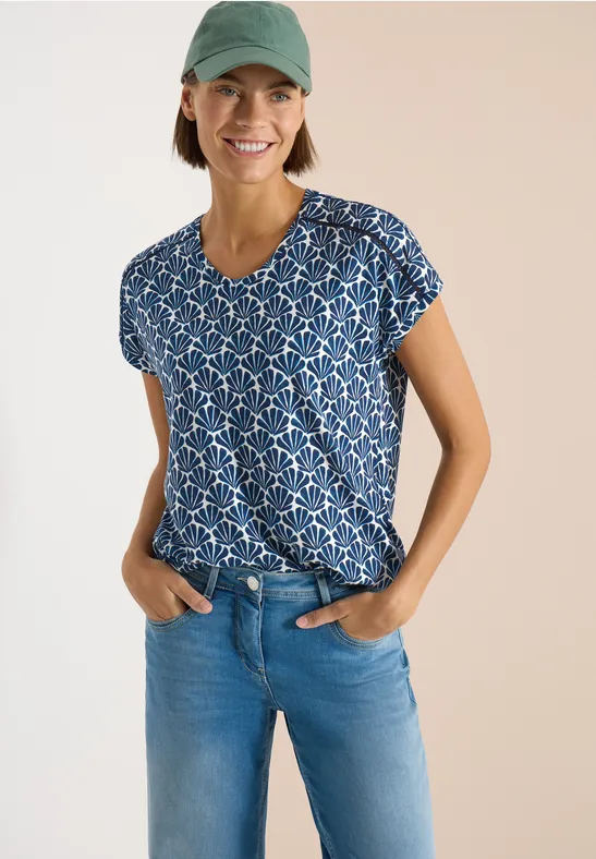 Person in blauem, gemustertem T-Shirt und hellblauen Jeans vor hellem Hintergrund.