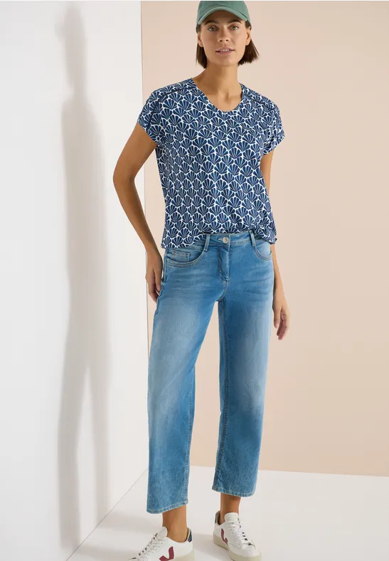 Frau trägt blaues Muster-T-Shirt, hellblaue gerade Jeans und weiße Sneaker vor neutralem Hintergrund.