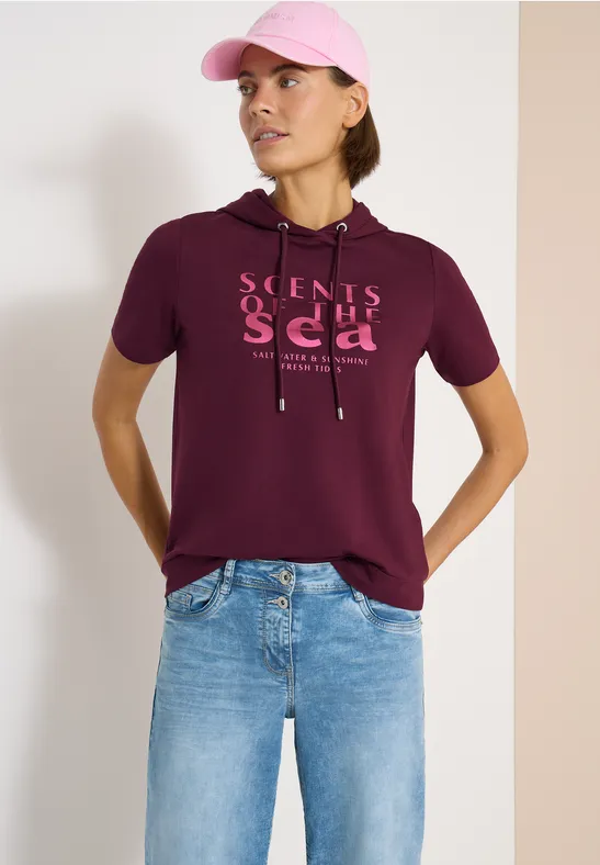 Frau trägt lila Kurzarm-Hoodie mit Aufschrift „Scenes of the sea“ und Jeans.