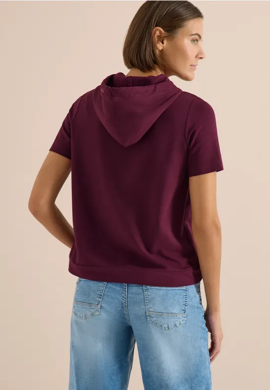 Frau von hinten in kurzem, bordeauxfarbenem Kapuzen-T-Shirt und Jeans.