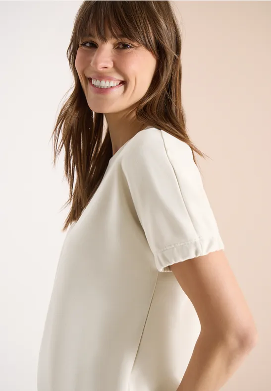 Frau in seitlicher Ansicht mit unscharfem Gesicht, trägt eine cremeweiße Bluse mit kurzen Ärmeln und eckigem Ausschnitt.