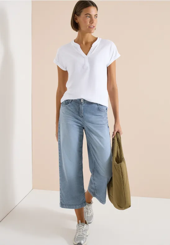 Frau in weißem Kurzarmshirt, hellen Culotte-Jeans und Sneakern mit grünem Shopper vor neutralem Hintergrund.