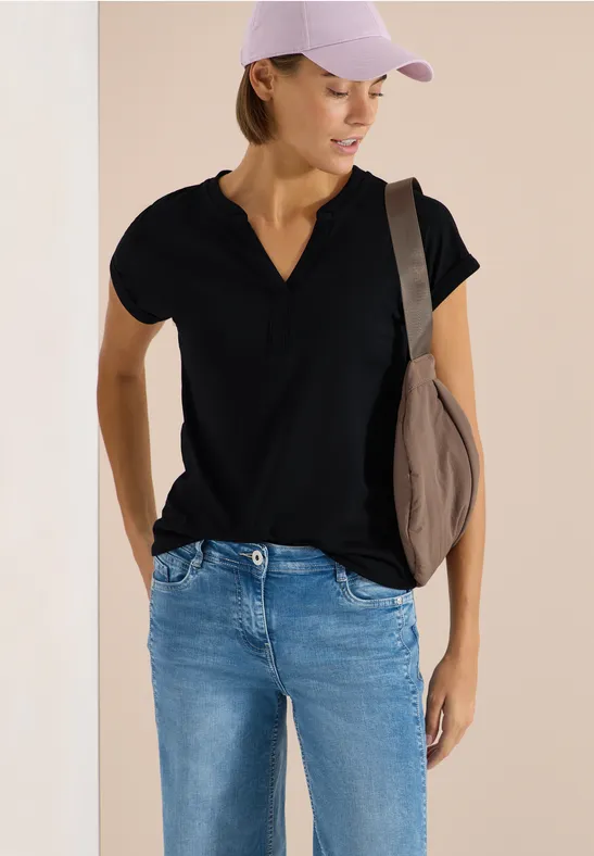 Frau in schwarzem V-Shirt, Jeans und Cap mit Umhängetasche vor hellem Hintergrund.