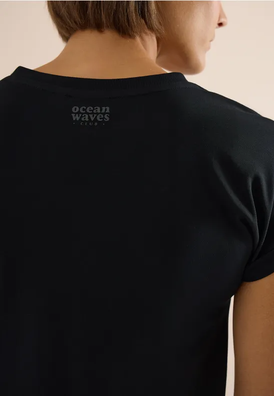 Schwarzes T-Shirt mit weißem Aufdruck 'ocean WAVES' auf dem Rücken, Person von hinten.