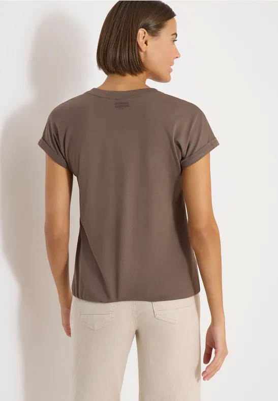 Frau von hinten in braunem T-Shirt und hellen Hosen vor hellem Hintergrund.