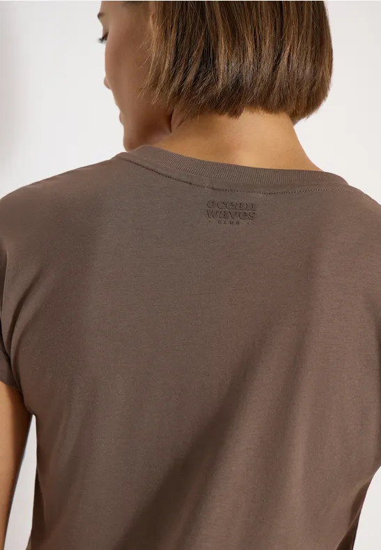 Rückansicht einer Frau mit kurzem braunen Haar, die ein schlichtes braunes T-Shirt mit kleinem Nackenprint trägt.