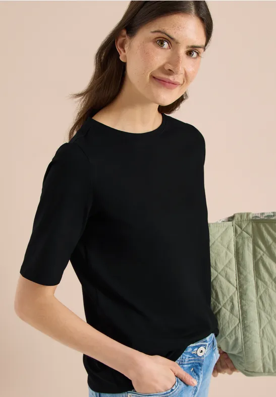 Frau in schwarzem T-Shirt und Jeans hält eine grüne Steppjacke über die Schulter.