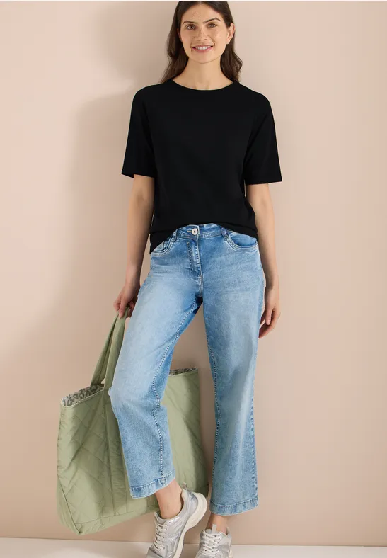 Frau in weitem Jeans, schwarzem T-Shirt und Sneakers trägt eine grüne Stofftasche vor beigem Hintergrund