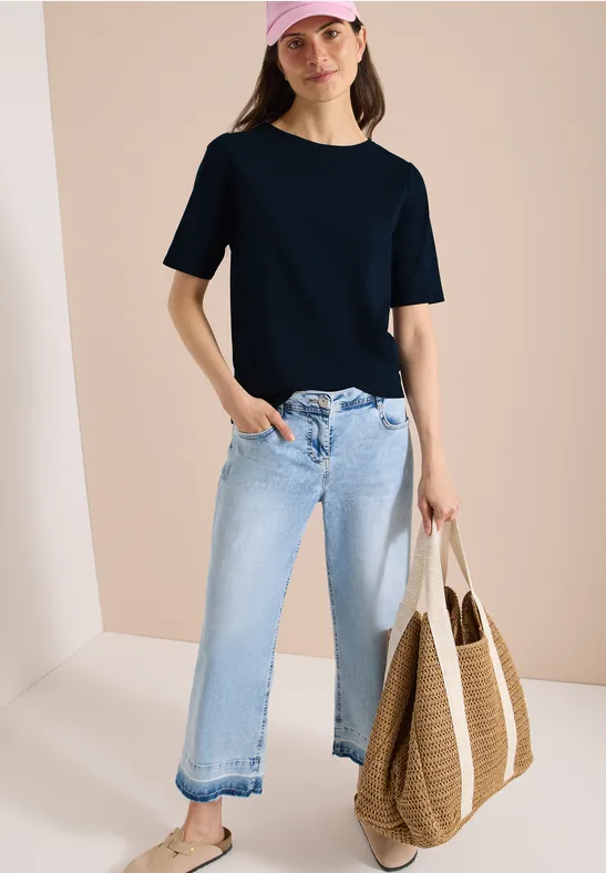 Frau in schwarzem T-Shirt, hellblauen weiten Jeans und Strohtasche vor hellem Hintergrund.
