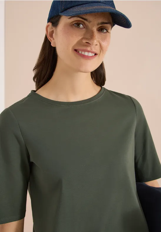 Person mit olivgrünem T-Shirt mit halblangen Ärmeln und Cap vor hellem Hintergrund.