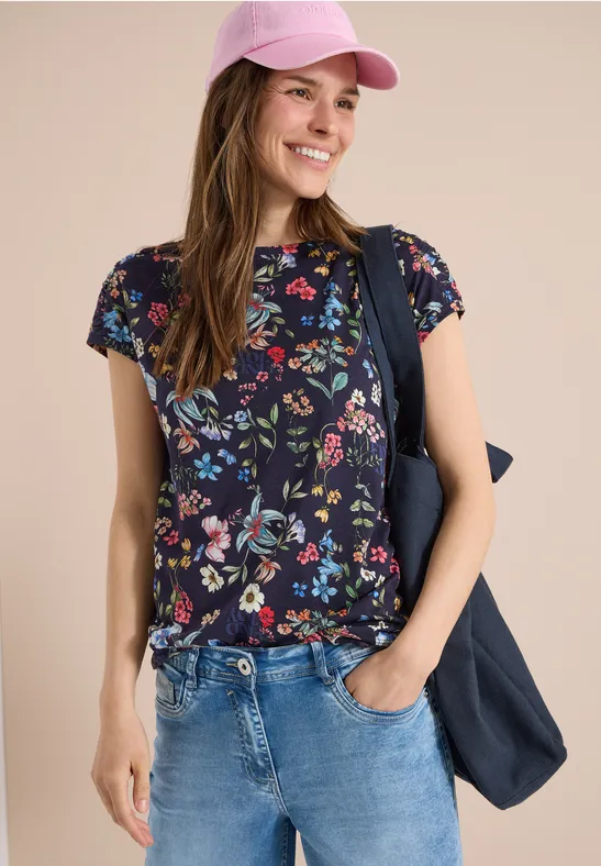 Person trägt Jeans, dunkles T-Shirt mit Blumenmuster, rosa Cap und blaue Tasche über der Schulter.