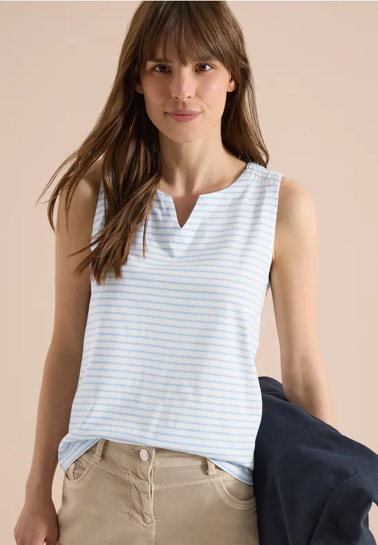 Femme en haut sans manches bleu clair à rayures blanches, pantalon clair et veste sombre à la main.