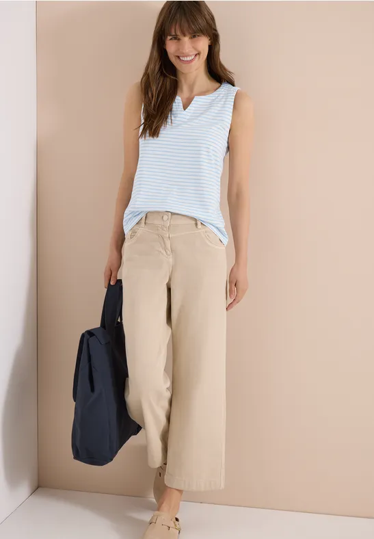 Femme en débardeur rayé bleu clair et pantalon large beige, tenant un sac bleu marine, posant devant un mur clair.