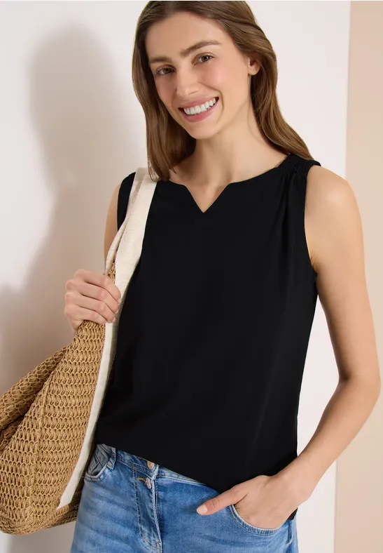 Femme en haut noir sans manches, tenant un panier en osier et posant une main dans la poche de son jean.