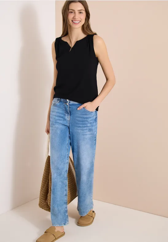 Femme en top noir et jean bleu devant un mur clair, portant un sac en osier et des mules.