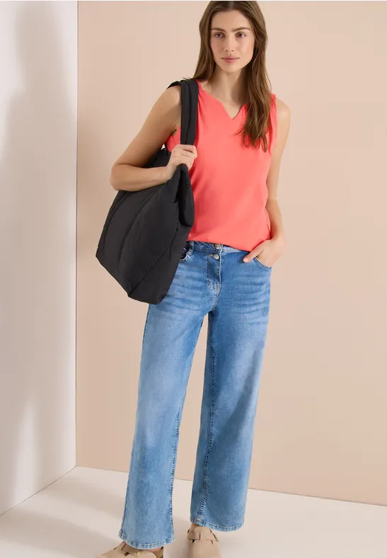 Frau mit rotem ärmellosem Top, blauen weiten Jeans und schwarzer Tasche vor neutralem Hintergrund.