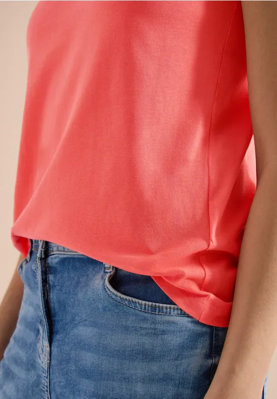 Eine Person trägt ein korallenrotes, ärmelloses Top, das in eine klassische blaue Jeans gesteckt ist.