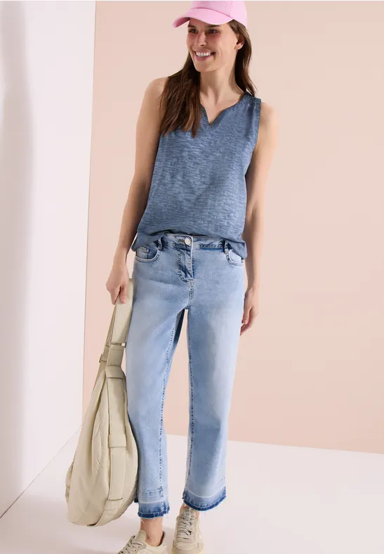 Eine Frau trägt ein ärmelloses, blaues Stricktop und helle Jeans, dazu eine beige Schultertasche.