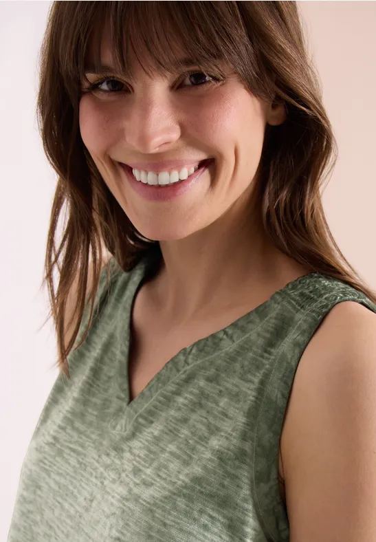 Person trägt ärmelloses grünes V-Ausschnitt-Oberteil, Gesicht unkenntlich, schulterlange Haare.