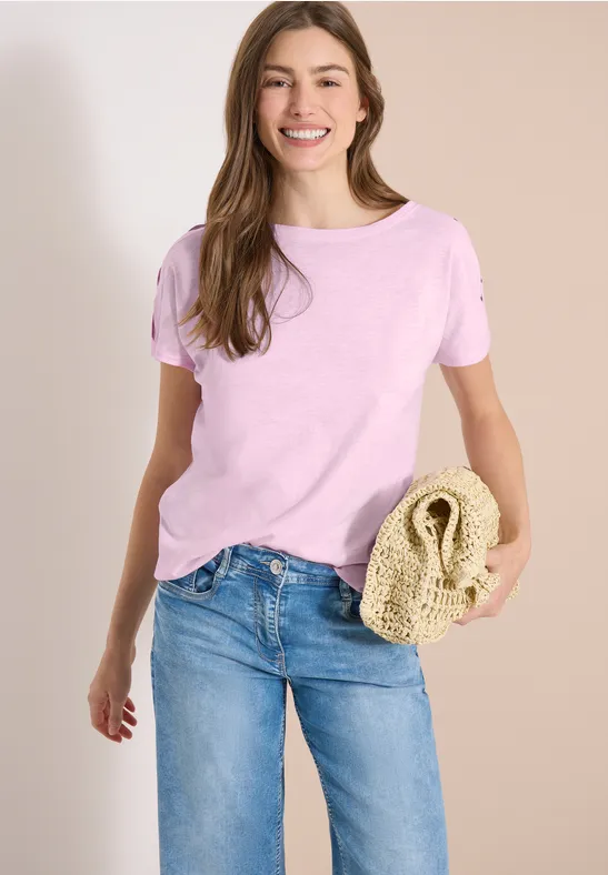 Femme en t-shirt rose et jean bleu tenant un sac en paille tressée devant un fond clair.