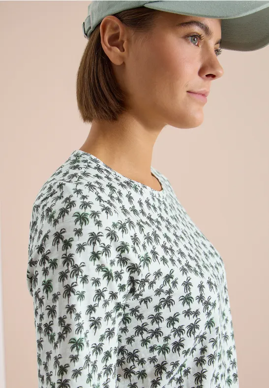 Seitliche Ansicht einer Frau mit Cap, die ein weißes T-Shirt mit grünen Palmenmuster trägt.