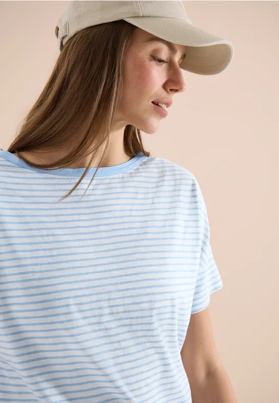 Person in gestreiftem T-Shirt und Baseballcap vor pastellfarbenem Hintergrund, Gesicht unscharf.