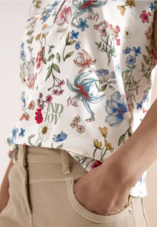 Tunique blanche à motif floral coloré, pantalon beige, main glissée dans la poche.