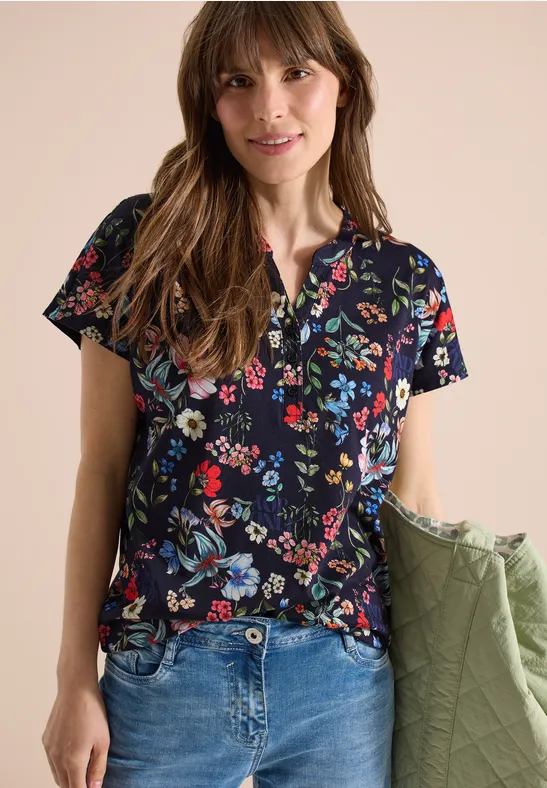 Frau trägt dunkelblaues, kurzärmeliges Shirt mit buntem Blumenmuster, blaue Jeans und hält grüne Steppjacke.