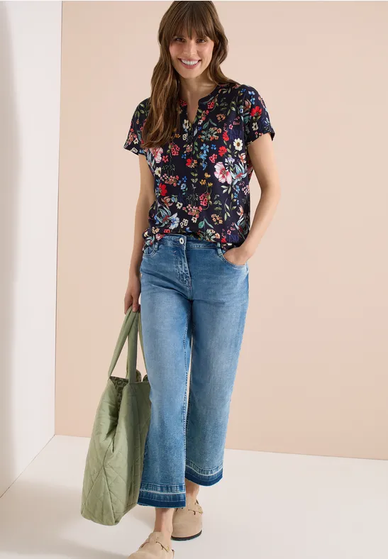 Frau in lässigem Blumen-Shirt, weiten Jeans, beigen Schuhen, grüner Tasche vor neutralem Hintergrund.