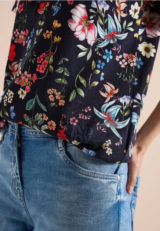 Person trägt blaue Jeans und schwarze Bluse mit buntem Blumenmuster, Hand in Tasche.