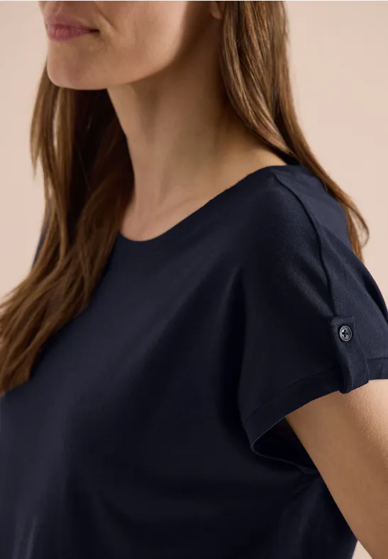 Femme aux cheveux bruns portant un T-shirt bleu marine à encolure bateau et bouton sur la manche.