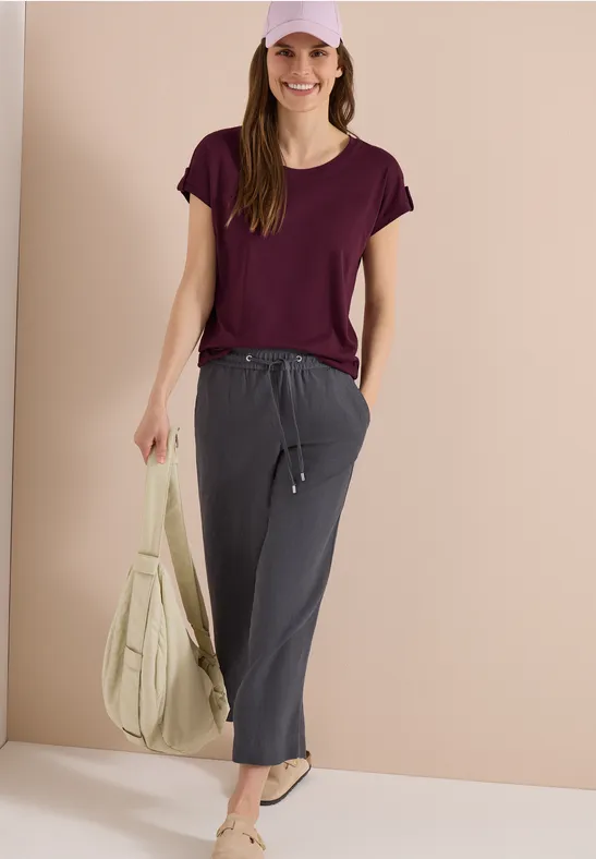 Femme en t-shirt violet, pantalon gris et sandales, tenant un sac beige devant un mur beige.