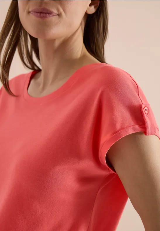 Eine Frau trägt ein korallenrotes Kurzarmshirt mit Knopfdetail am Ärmel vor neutralem Hintergrund.