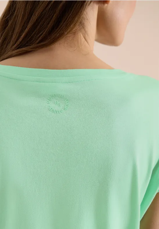 Nahaufnahme des Rückens einer Frau in mintgrünem T-Shirt vor neutralem Hintergrund.