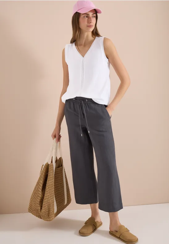 Model mit weißen V-Ausschnitt-Top, lockeren grauen Culottes, Basttasche, beigen Slippern und rosa Cap.