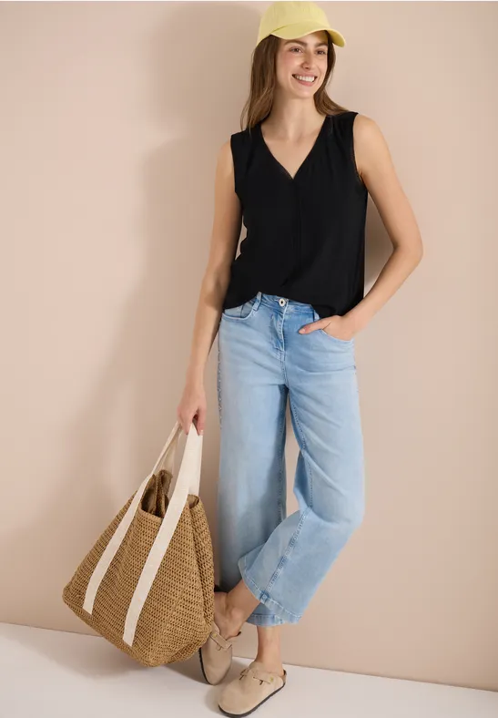 Frau trägt schwarzes Top, hellblaue Jeans, Loafers und hält eine Strohtasche vor beigem Hintergrund.