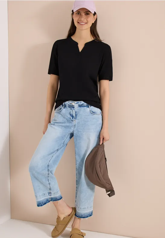 Person lehnt an Wand, trägt schwarze Bluse, weite hellblaue Jeans, senfgelbe Schuhe, hält braune Jacke.