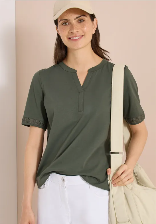 Eine Frau in dunkelgrünem Henley-Shirt mit kurzen Ärmeln und beiger Umhängetasche vor hellem Hintergrund.